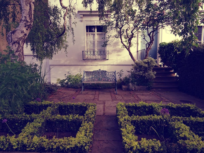 A small front garden featuring a paved patio area with a decorative metal bench positioned against the white exterior wall of a house, which has a window with detailed white trim and a small balcony. 