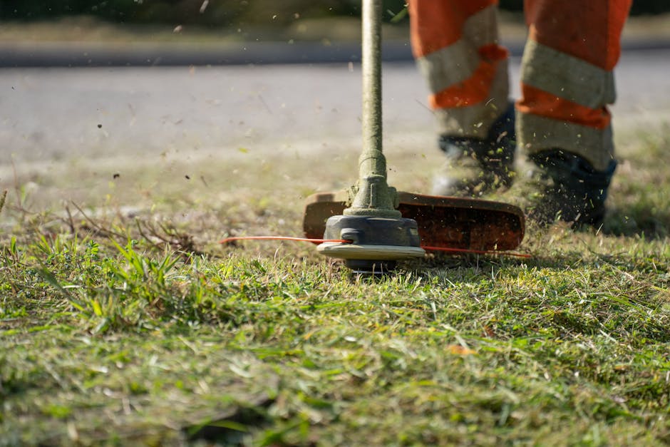 A person dressed in a blue waterproof jacket with the hood up and black outdoor trousers is engaging in garden maintenance, using a metal rake to tidy up fallen leaves and debris on the vibrant, neatl